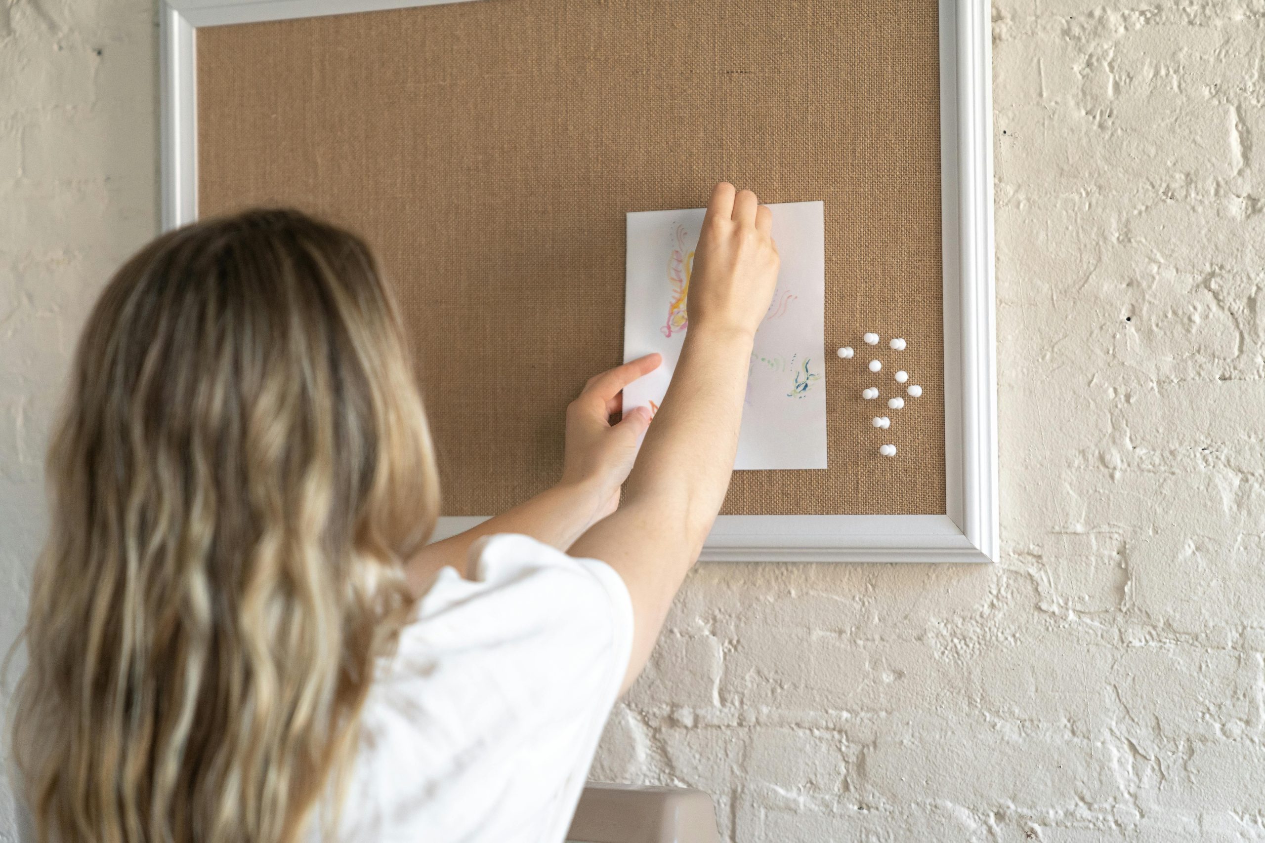 Blonde woman attaching a drawing to a cork board in an office setting, creating an inspiring workspace.